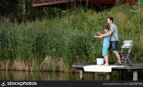 Positive father helping his teenage boy to fight a fish they caught. They are spinning fishing reel while fishing together from wooden pier on the pond. Cute son and dad catching fish with fishing rod and reel while spending leisure on the lake.