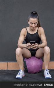 Positive female athlete in sportswear with ponytail sitting on ball against black wall and browsing cellphone during break in fitness workout in gym. Sportswoman using smartphone during break