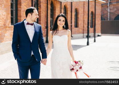 Positive female brunette in white wedding dress, holds bouquet and bridegroom`s hand, celebrate their wedding together, wait for guests. Happy married couple became newlyweds. Relationship concept