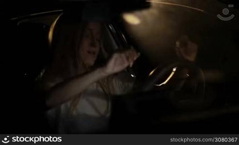 Positive female driver sitting in modern car listening to music and making dancing moves while enjoying leisure at night. Beautiful blonde woman enjoying her favorite song on the radio and dancing while sitting in parked car on a summer night.