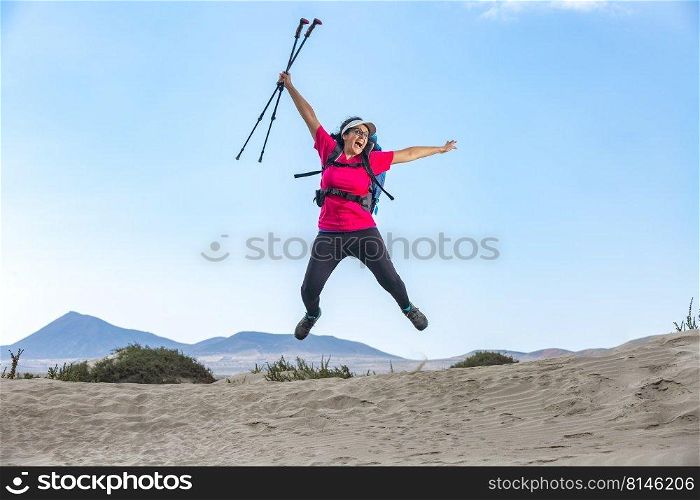 Positive female explorer with backpack and trekking poles jumping above sandy Famara beach and celebrating achievement. Excited traveler jumping in highlands