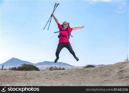 Positive female explorer with backpack and trekking poles jumping above sandy Famara beach and celebrating achievement. Excited traveler jumping in highlands