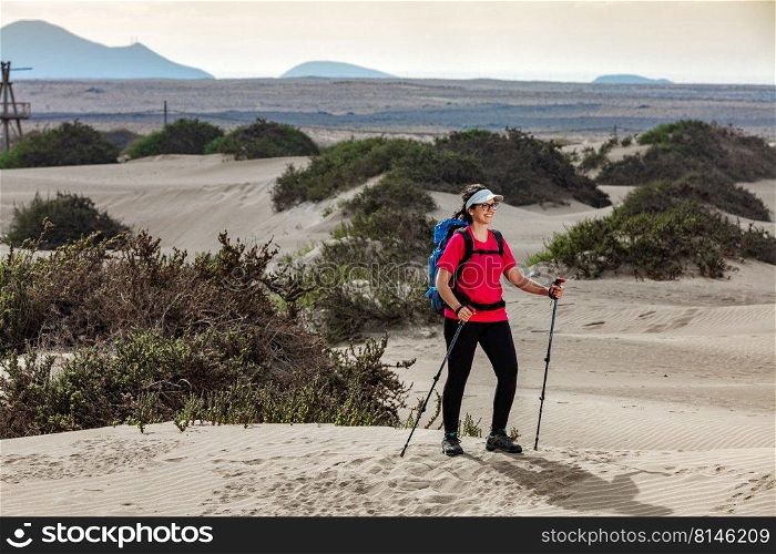 Positive female hiker with trekking sticks and backpack standing on Famara shore in summer and looking away. Traveling woman with trekking poles on sandy shore