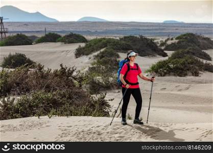 Positive female hiker with trekking sticks and backpack standing on Famara shore in summer and looking away. Traveling woman with trekking poles on sandy shore