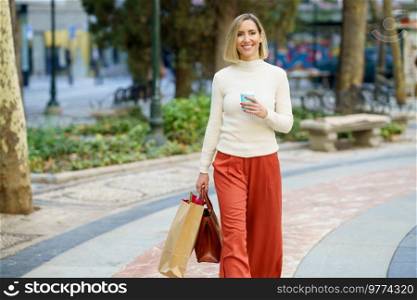 Positive female in casual clothes carrying shopping bag and handbag and having cup of takeaway beverage in park. Smiling woman with shopping bags and takeaway drink