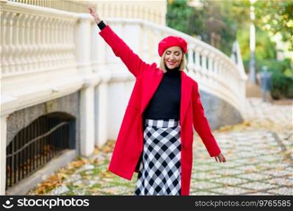 Positive female in red clothes with beret strolling with raised arm and closed eyes on street along white fence in city. Cheerful woman in stylish wear walking near fence
