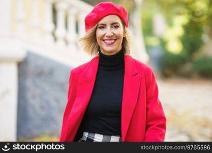 Positive female in trendy red outfit looking at camera with flying hair while strolling in street of city against blurred background. Happy woman in beret walking on street