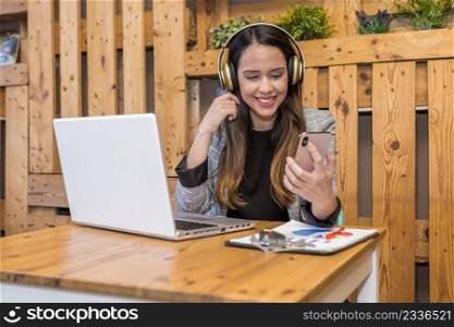 Positive female remote worker in wireless headphones sitting at wooden table and checking messages on cellphone during online work on netbook. Smiling woman reading message on smartphone while working on laptop