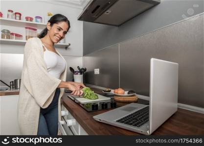 Positive female standing at counter and cutting lettuce while watching recipe on netbook and cooking lunch. Content woman watching video on laptop and cooking in kitchen