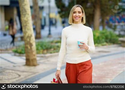 Positive female wearing turtleneck and pants walking with bag and holding cup of takeaway coffee drink in park. Smiling woman with cup of beverage carrying paper bag