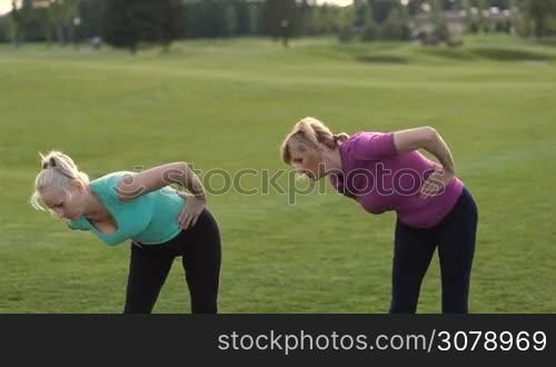Positive fitness adult women in sports clothes doing stretching exercises with trunk bending on park lawn. Two sporty senior females working out, doing tilts around and physical exercises during training on fresh air.