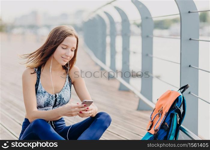 Positive fitness girl in top and leggings, has glad expression as reads pleasant text message recieved from boyfriend, has dark hair, rests outdoor. People, sport, modern technologies and lifestyle