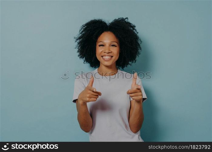 Positive friendly dark skinned lady making finger gun gesture and smiling at camera, happy young african female in white tshirt being happy to meet best friend, isolated over blue background. Positive friendly dark skinned lady making finger gun gesture while standing on blue background
