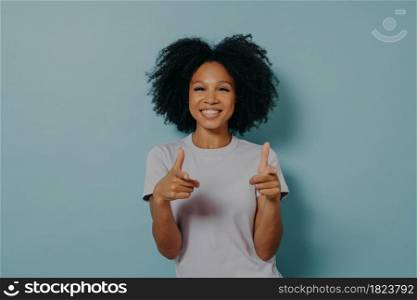 Positive friendly dark skinned lady making finger gun gesture and smiling at camera, happy young african female in white tshirt being happy to meet best friend, isolated over blue background. Positive friendly dark skinned lady making finger gun gesture while standing on blue background