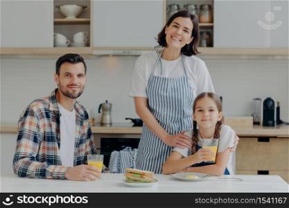 Positive friendly father, mother and their small daughter being in good mood, spend time together, pose at kitchen, gather to have breakfast, enjoy domestic atmosphere and day off. Time to eat