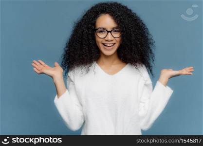Positive friendly young woman with Afro appearance, spreads palms, holds invisible object, has charming smile, healthy skin, wears spectacles and white sweater, isolated over blue background.