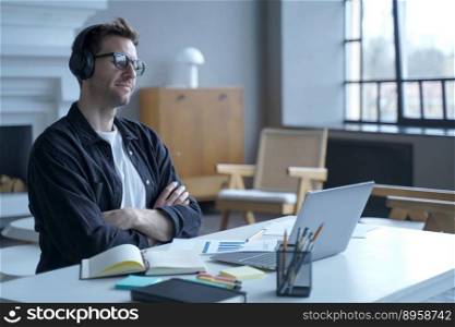 Positive German man bank employee sitting at workplace at modern home office in wireless headphones while using laptop working remotely online, looking at screen listening to distance learning courses. Positive German man bank employee sitting at workplace at modern home office in wireless headphones