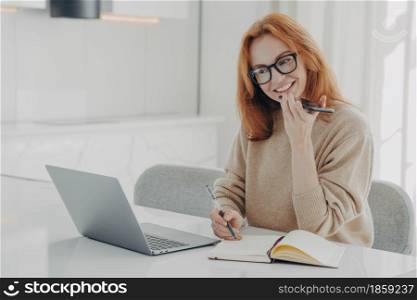 Positive ginger woman in casual clothes records audio message or smartphone, talks with virtual digital voice assistant while working on laptop, sits at her workplace, female uses speakerphone at work. Positive ginger woman in casual clothes records audio message or smartphone while working on laptop