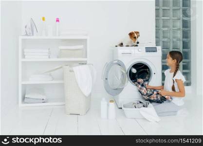 Positive girl emptying washing machine, holds clean checkered shirt, looks with smile at favourite pet who helps with doing laundry, poses on white floor with basin full of clothes, cleaning agents.