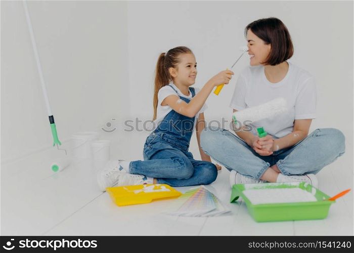 Positive girl smearks mums nose with paint by roller, have fun while making house renovation, sit together on floor in white room, use necessary tools for repairment. House renovation concept