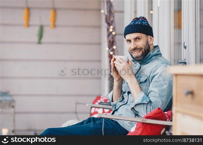 Positive glad attractive male model holds cup of tea, has good rest at balcony, breathes fresh air and enjoys sunny weather, has pleasant smile on face, feels delighted and pleased. People and drinks