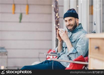 Positive glad attractive male model holds cup of tea, has good rest at balcony, breathes fresh air and enjoys sunny weather, has pleasant smile on face, feels delighted and pleased. People and drinks