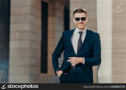 Positive glad young male wears formal suit, wears trendy shades, keeps hand in pocket, stands against office interior, being in good mood after successful business meeting. Leadership concept