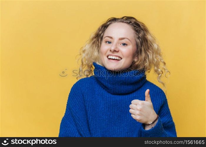 Positive good looking female model being satisfied with something, raises thumb, has glad expression, wears casual blue sweater, poses against yellow studio background. Body language concept