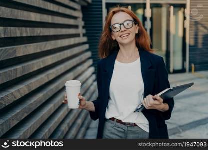 Positive good looking redhead woman enjoys walk outdoor drinks takeaway coffee carries digital electronic device and notepad wears formal clothes returns from work has excellent mood. Lifestyle. Redhead woman enjoys walk outdoor drinks takeaway coffee carries digital electronic device notepad