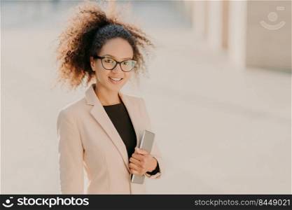 Positive gorgeous Afro American businesswoman strolls outdoor with touchpad, wears formal clothes, optical spectacles, smiles at camera, enjoys spare time after meeting. People and technology concept