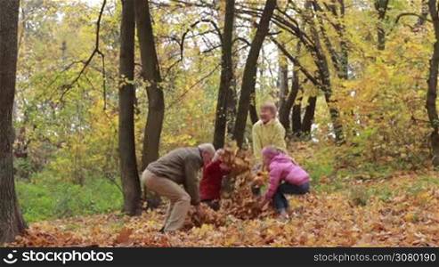 Positive grandparents and smiling grandchildren throwing leaves in autumn park. Cheerful senior couple and adorable sibling playing in public park on a warm fall day. Multi generation family spending leisure outdoors in autumn time. Slow motion.