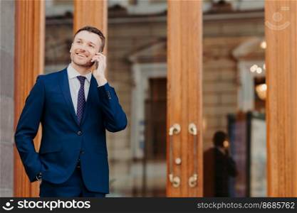 Positive handsome male consults with bank operator about transactions via cell phone, dressed in formal suit, keeps hand in pocket, smiles positively, stands near door or entrance of restaurant