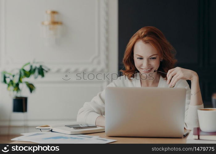 Positive happy female freelancer has busy working day, works distantly from home, sits in front of laptop computer against modern interior, works on creative task, watches webinar for improving skills