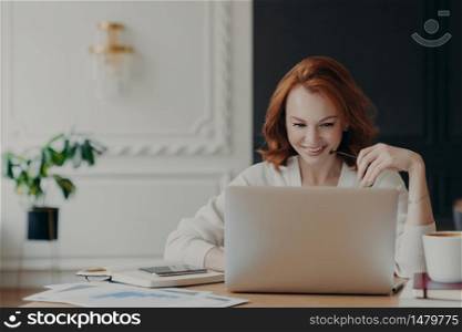 Positive happy female freelancer has busy working day, works distantly from home, sits in front of laptop computer against modern interior, works on creative task, watches webinar for improving skills