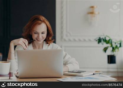 Positive happy female freelancer has busy working day, works distantly from home, sits in front of laptop computer against modern interior, works on creative task, watches webinar for improving skills