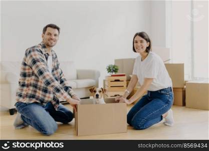 Positive happy husband and wife stand on knees near carton box with cute dog, enjoy relocation in new apartment, smiles pleasantly at camera. Pedigree pet rides in cardboard container on floor