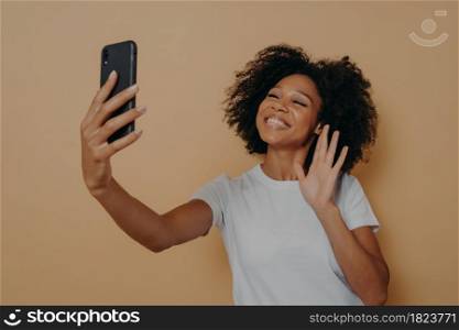 Positive happy mixed race woman waving with hand at camera during video call with friend on modern smartphone, gesturing hello to her followers in social media while posing isolated over studio wall. Positive happy mixed race woman waving with hand at camera during video call on modern smartphone
