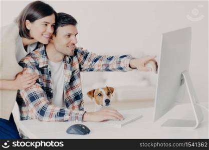 Positive happy young woman and man work remotely at computer, spend leisure together, indicates in monitor, makes booking online, search information about hotels, make planning for future travel