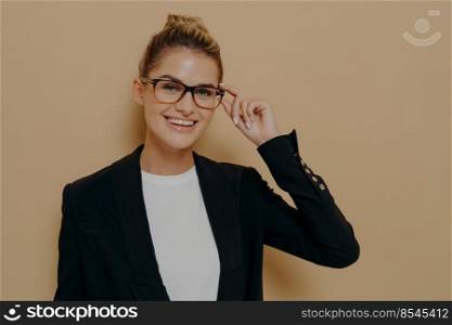 Positive happy young woman touching rim of glasses and looking through them, smiling female student touching spectacles while looking at camera with smile, isolated over beige wall with copy space. Positive happy young woman touching rim of glasses and looking through them