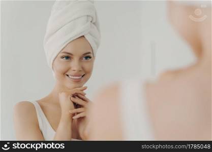 Positive healthy young woman smiling while applying facial cream reflecting in mirror, happy attractive lady putting moisturizing nourishing cream during morning beauty routine in bathroom. Healthy young woman with clean skin smiling while applying facial cream