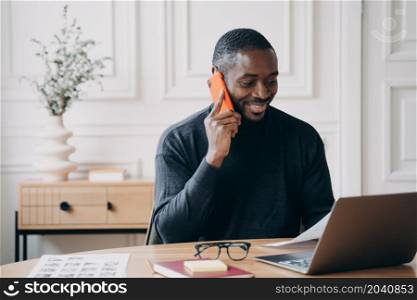 Positive joyful Afro american male developer sitting at desk looking at computer screen, speaking on smartphone with customer, discuss application working from home solve business issues distantly. Joyful Aframerican male developer sits at desk looks at computer screen while speaking on smartphone