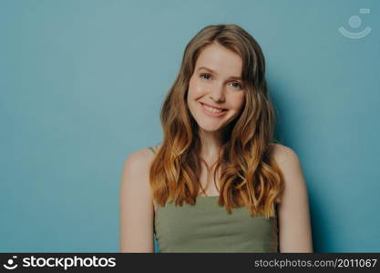 Positive joyful teen girl in top tank demonstrating optimism and happiness while standing isolated over blue studio background with copy space for text, pleased happy female looking at camera. Positive joyful teen girl in top tank demonstrating optimism and happiness, isolated over blue wall
