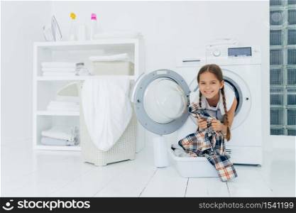 Positive kid with pigtails sticks out head from washing machine, has fun and holds shirt, prepares for washing, smiles gently, spends free time in laundry room. Children, cleanliness, hygiene concept