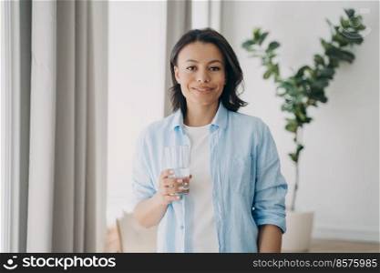 Positive lady is holding glass of pure water. Pretty caucasian woman is standing in room of apartment. Concept of daily health care, hydration and nutrition. Healthy lifestyle, wellbeing and wellness.. Pretty caucasian woman is holding glass of pure water. Daily health care, hydration and nutrition.