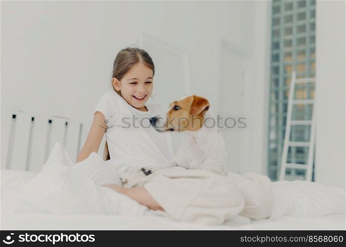 Positive little girl plays with pedigree pet, dressed in white pyjamas, spends leisure time in bed, has fun at bedroom smiles happily. Dog poses near its small kid owner. Children, animals and leisure