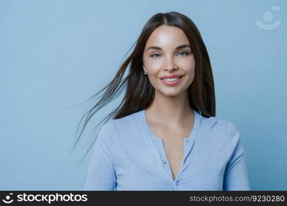 Positive lovely feminine lady happy to have first date with boyfriend, smiles broadly, has appealing appearance, glad to hear something pleasant, models indoor alone, wears blue casual jumper