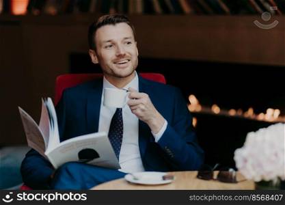 Positive male company owner recreats in cozy restaurant, holds white cup with cappuccino, has pleasant smile on face, reads business journal, notices beautiful woman aside, has spare time after work