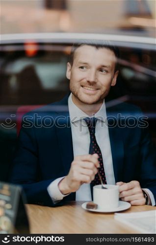 Positive male entrepreneur in formal clothing, drinks coffee, has cheerful expression, looks happily aside, spends free time in cafe, poses near window. People, business meeting and rest concept