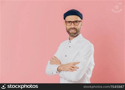 Positive male entrepreneur with self confident satisfied facial expression, keeps hands crossed, stands in profile, looks directly at camera, poses indoor against pink background with free space