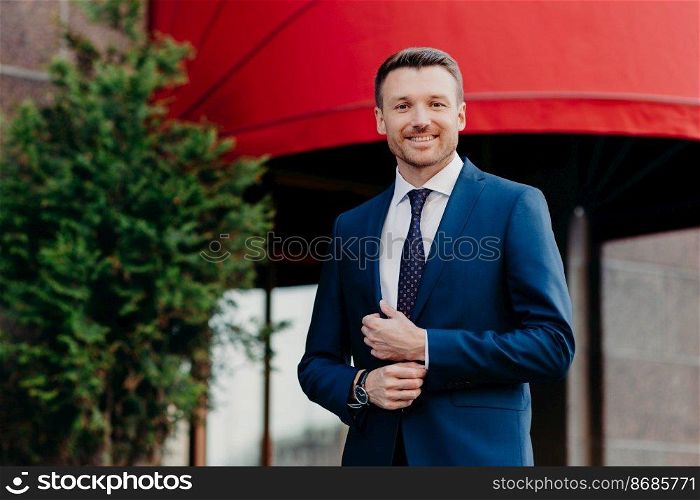 Positive male executive worker wears formal black suit, has positive expression, stands outdoor, waits for someone, being confident in achieving great success during his hard work and persistance